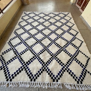 May include: A white rug with a black and blue diamond pattern. The rug has a fringe border.