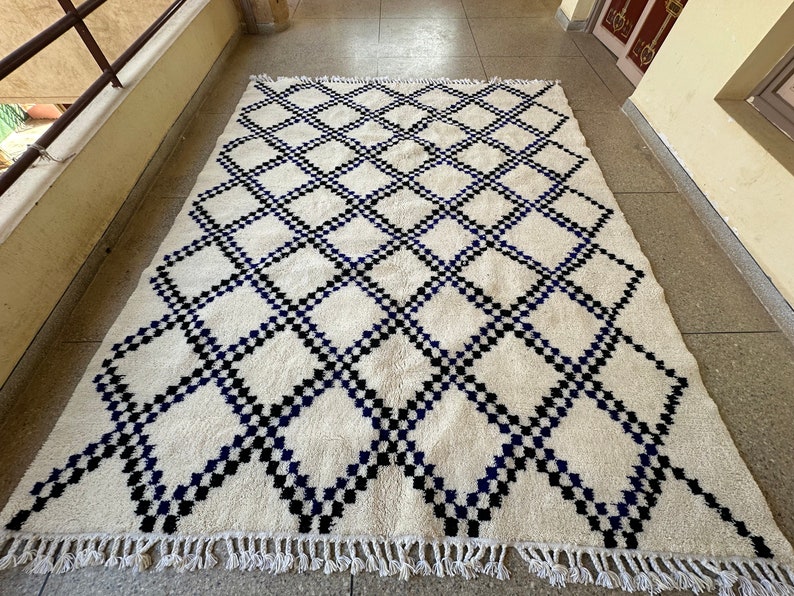 May include: A white rug with a black and blue diamond pattern. The rug has a fringe border.