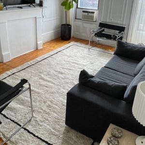 May include: A black couch with a white rug in a living room. The couch has a black and white patterned throw pillow. There is a black chair with a chrome frame in the foreground.