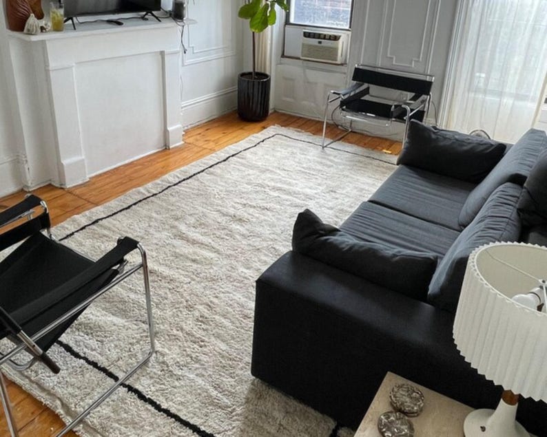 May include: A black couch with a white rug in a living room. The couch has a black and white patterned throw pillow. There is a black chair with a chrome frame in the foreground.