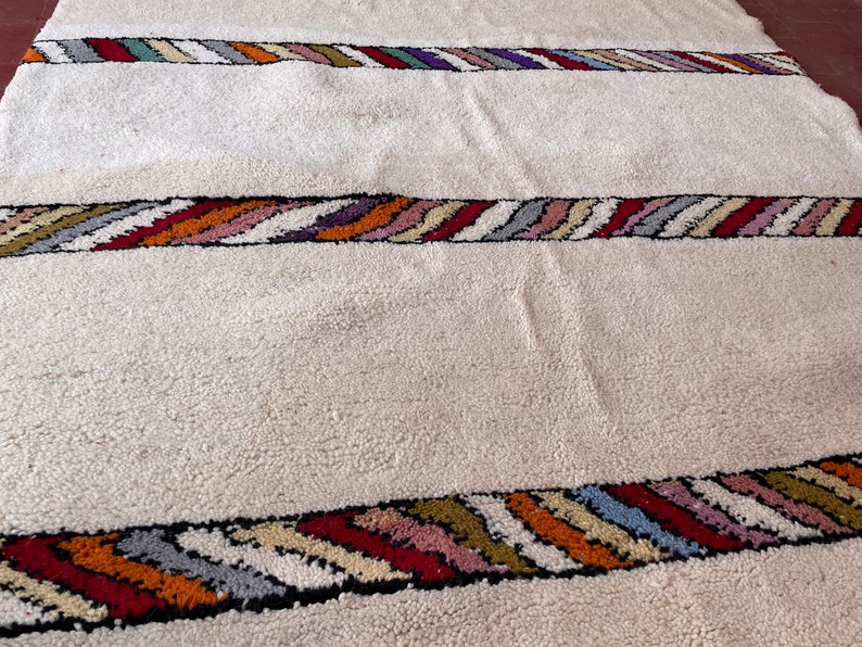 Puede incluir: Una alfombra blanca con rayas onduladas de colores a lo largo de los bordes. Las rayas est&aacute;n hechas de diferentes colores, incluyendo rojo, naranja, amarillo, verde, azul y morado.