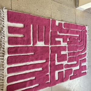 May include: A rectangular rug with a geometric design in shades of pink and white. The rug features a textured, raised pattern in pink against a white background, with fringed edges. The design is abstract and modern.