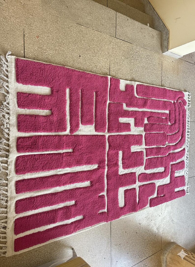 May include: A rectangular rug with a geometric design in shades of pink and white. The rug features a textured, raised pattern in pink against a white background, with fringed edges. The design is abstract and modern.