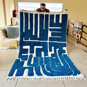 May include: A large, rectangular rug with a bold, abstract design in shades of blue and white. The rug features a geometric pattern with thick, textured lines and a fringed edge. The rug is being held up by a person.