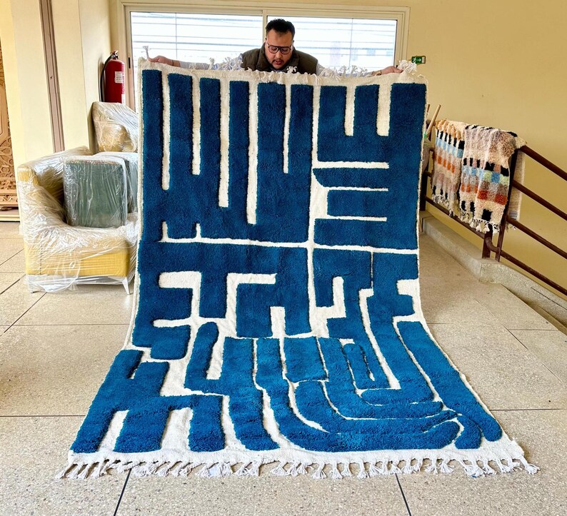 May include: A large, rectangular rug with a bold, abstract design in shades of blue and white. The rug features a geometric pattern with thick, textured lines and a fringed edge. The rug is being held up by a person.