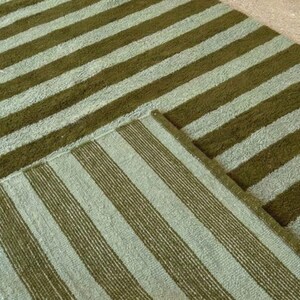 May include: Two rectangular rugs with a striped pattern. The stripes alternate between a light green and a darker olive green. The rugs are made of a textured material and are placed on a light-colored surface.