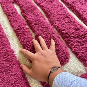 May include: A close-up view of a rug with a textured, plush design. The rug features alternating stripes of fuchsia and off-white. The fuchsia stripes have a raised, looped texture. A hand wearing a bracelet is resting on the rug.