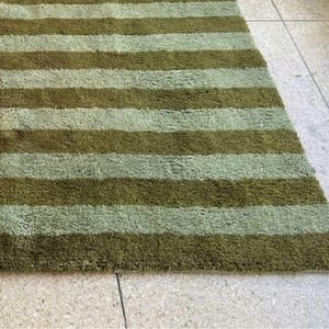 May include: A rectangular rug with alternating horizontal stripes of olive green and light green. The rug has a shaggy texture and is placed on a light-colored tiled floor. The rug's design is simple and modern, suitable for various interior styles.