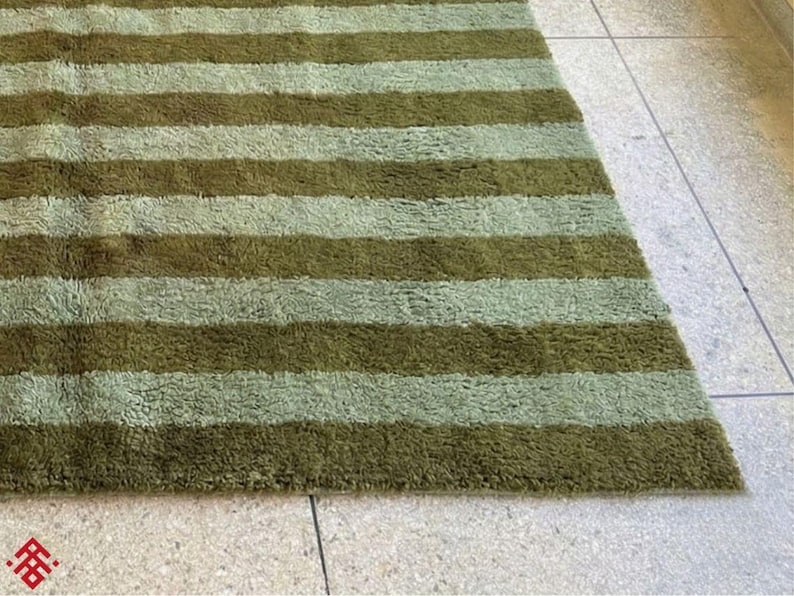 May include: A rectangular rug with alternating horizontal stripes of olive green and light green. The rug has a shaggy texture and is placed on a light-colored tiled floor. The rug's design is simple and modern, suitable for various interior styles.