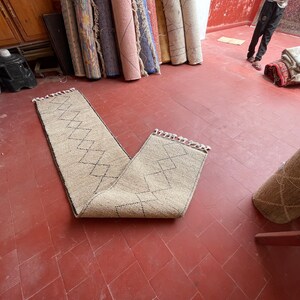 May include: A beige runner rug with a diamond pattern in black. The rug is folded in half and laid out on a red tiled floor.