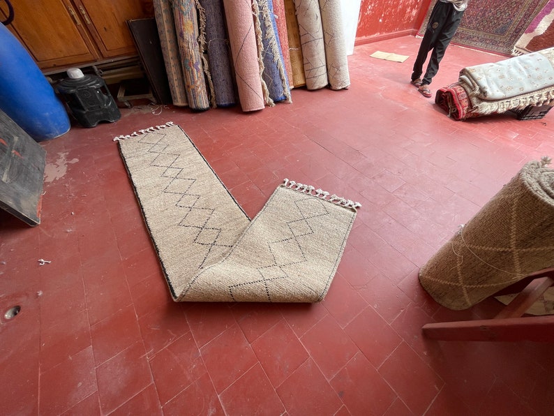 May include: A beige runner rug with a diamond pattern in black. The rug is folded in half and laid out on a red tiled floor.