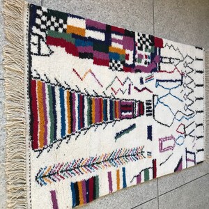 May include: A white rug with a colorful geometric pattern. The rug has a fringe border and features a variety of shapes and colors, including red, blue, green, yellow, and purple.