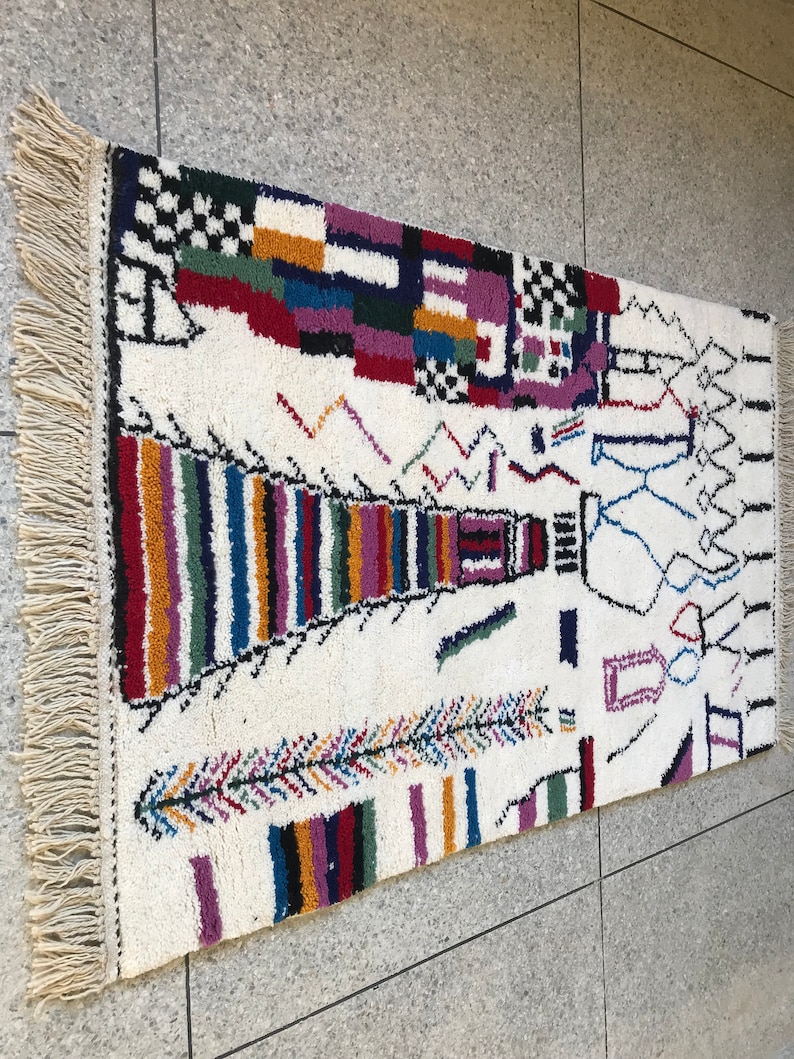 May include: A white rug with a colorful geometric pattern. The rug has a fringe border and features a variety of shapes and colors, including red, blue, green, yellow, and purple.