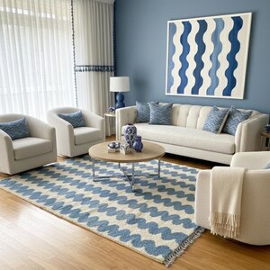 Pu&ograve; includere: Un soggiorno con uno schema di colori blu e bianco. La stanza presenta un divano bianco, poltrone e un tappeto con un motivo a onde blu. Un grande dipinto con disegni di onde blu &egrave; appeso alla parete blu. Un tavolino rotondo in legno &egrave; al centro.