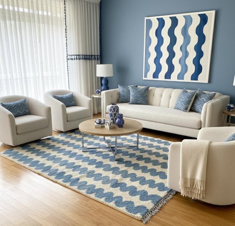 Pu&ograve; includere: Un soggiorno con uno schema di colori blu e bianco. La stanza presenta un divano bianco, poltrone e un tappeto con un motivo a onde blu. Un grande dipinto con disegni di onde blu &egrave; appeso alla parete blu. Un tavolino rotondo in legno &egrave; al centro.
