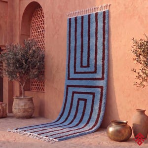 Peut inclure: Un long tapis rectangulaire présentant une base bleu clair avec des motifs géométriques en forme de U marron foncé. Le tapis possède un poil épais et texturé, terminé par des franges nouées blanches sur les bords supérieur et inférieur.