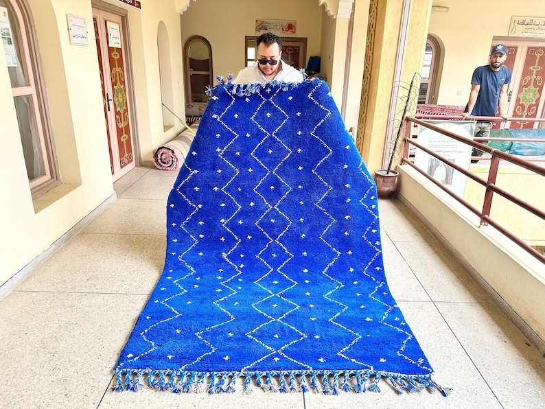 Puede incluir: Una alfombra marroqu&iacute; azul y blanca con un patr&oacute;n de diamantes. La alfombra est&aacute; doblada por la mitad y la est&aacute; sujetando una persona.