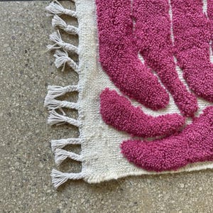 May include: A close-up of a textured rug with a cream-colored base and a pattern of magenta shapes. The rug has a fringe of tassels along one edge. The rug is on a speckled gray surface.