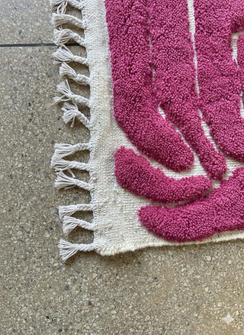 May include: A close-up of a textured rug with a cream-colored base and a pattern of magenta shapes. The rug has a fringe of tassels along one edge. The rug is on a speckled gray surface.