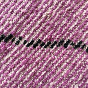 May include: Close-up of a woven fabric with a diagonal pattern in shades of pink and white. A black stripe runs horizontally across the fabric.