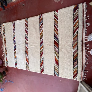 Puede incluir: Una alfombra blanca con rayas de colores tejidas en la tela. Las rayas est&aacute;n hechas de hilos rojos, naranjas, amarillos, azules y verdes. La alfombra tiene un borde de flecos.