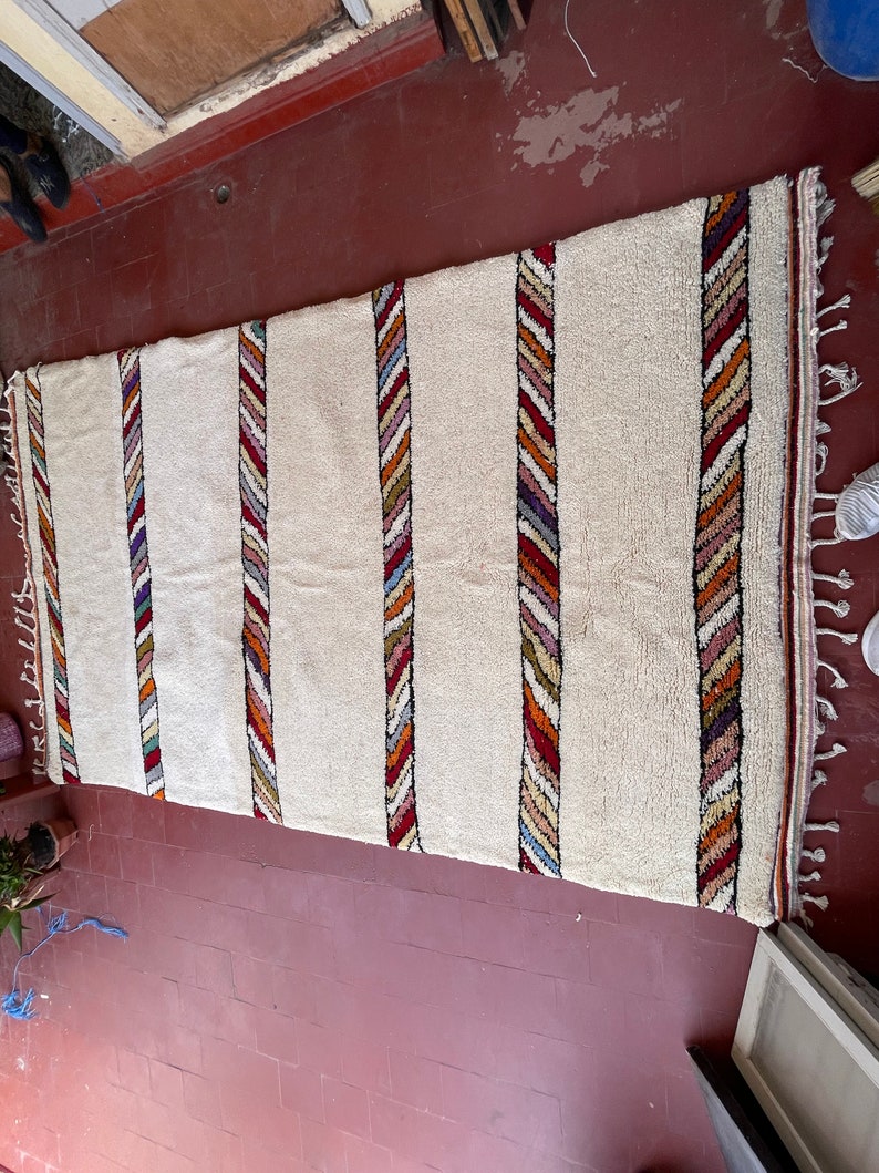 Puede incluir: Una alfombra blanca con rayas de colores tejidas en la tela. Las rayas est&aacute;n hechas de hilos rojos, naranjas, amarillos, azules y verdes. La alfombra tiene un borde de flecos.