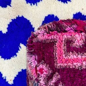 Pu&ograve; includere: Primo piano di un tappeto vibrante e strutturato con un motivo geometrico. Il tappeto presenta uno sfondo blu intenso con forme a zig-zag bianche. Una sezione di un tappeto rosa e viola con un disegno floreale &egrave; in primo piano.