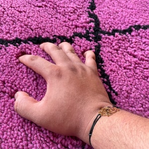 May include: A hand resting on a pink and black patterned rug. The hand is wearing a black string bracelet with a gold charm.