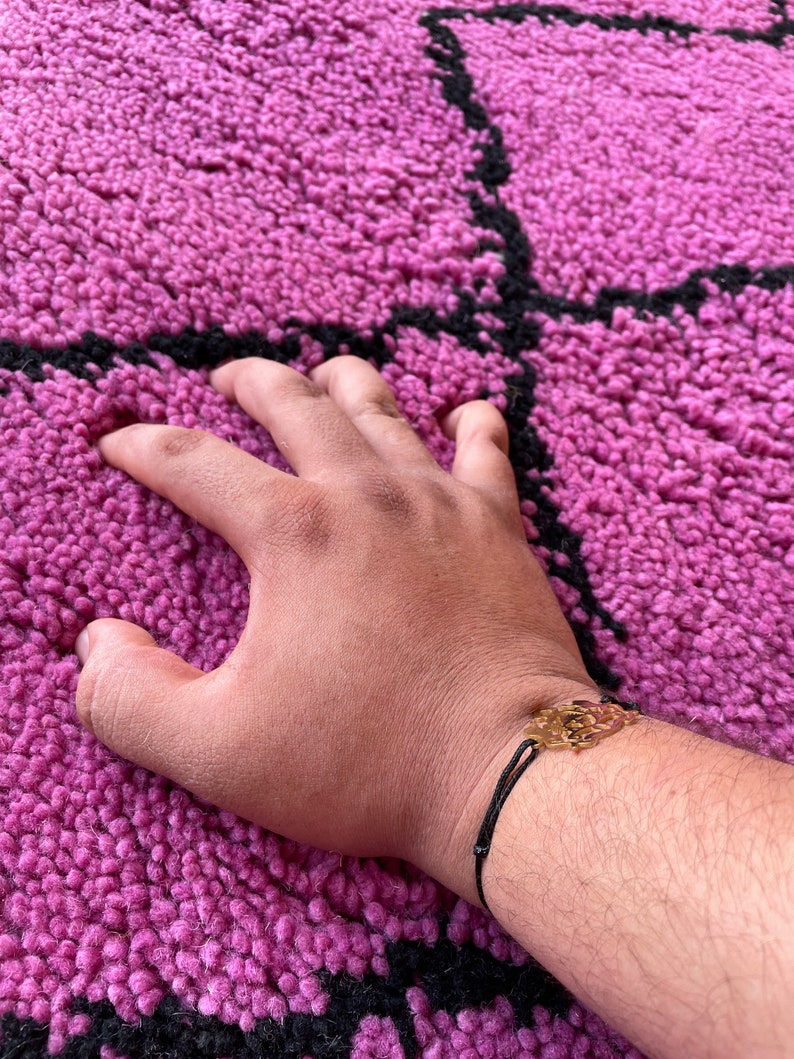 May include: A hand resting on a pink and black patterned rug. The hand is wearing a black string bracelet with a gold charm.