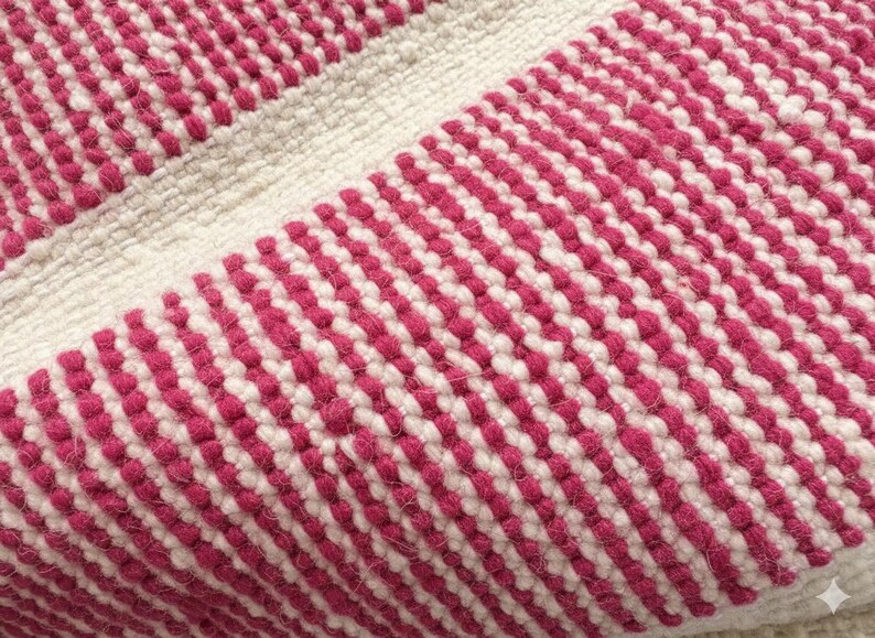 May include: Close-up of a textured rug with a repeating pattern of alternating white and magenta stripes. The rug appears to be made of woven material, with a soft, tactile appearance. The design is simple yet visually appealing.