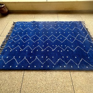 Puede incluir: Una alfombra azul y blanca con un dise&ntilde;o geom&eacute;trico. La alfombra tiene un borde de flecos y est&aacute; hecha de un material suave y mullido.