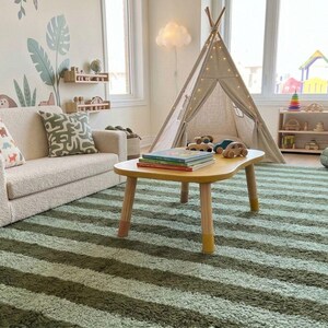 May include: A children's playroom with a cream-colored sofa, a yellow table, and a striped green rug. A canvas teepee is in the background, along with wooden toys and a cactus. The walls are decorated with jungle-themed murals.