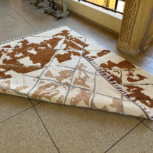 May include: A white and brown patterned rug with fringe on the edges. The rug is folded in half and lying on a tiled floor.
