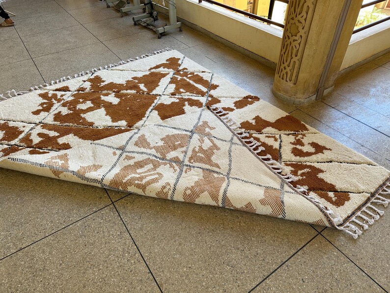 May include: A white and brown patterned rug with fringe on the edges. The rug is folded in half and lying on a tiled floor.