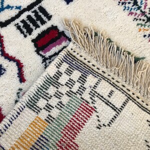 May include: A white rug with colorful geometric patterns and fringe. The rug has black, red, blue, green, yellow, and white stripes.