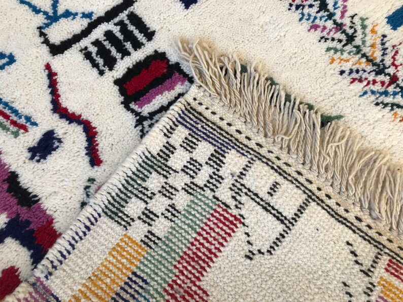 May include: A white rug with colorful geometric patterns and fringe. The rug has black, red, blue, green, yellow, and white stripes.
