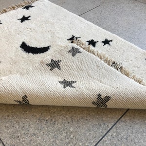 May include: A white rug with a black crescent moon and stars design. The rug has a fringed edge and is folded in half.