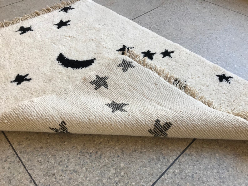 May include: A white rug with a black crescent moon and stars design. The rug has a fringed edge and is folded in half.