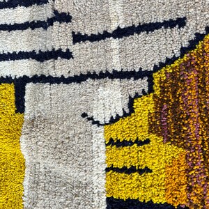 Puede incluir: Primer plano de una alfombra abstracta colorida con formas geom&eacute;tricas en tonos de amarillo, marr&oacute;n, negro y blanco. La alfombra est&aacute; hecha de un material grueso y texturizado.