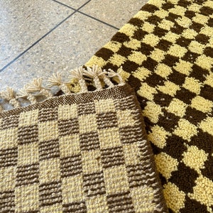 May include: Two woven rugs with a checkered pattern in brown and cream. The rug on the left has a fringe border.