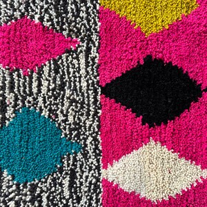 K&ouml;nnte beinhalten: Nahaufnahme eines bunten Teppichs mit geometrischem Muster. Der Teppich besteht aus einem weichen, flauschigen Material und zeigt eine Vielzahl von Farben, darunter Pink, Schwarz, Wei&szlig;, T&uuml;rkis und Gelb.