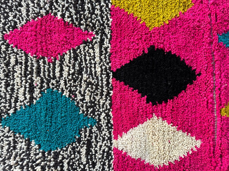 K&ouml;nnte beinhalten: Nahaufnahme eines bunten Teppichs mit geometrischem Muster. Der Teppich besteht aus einem weichen, flauschigen Material und zeigt eine Vielzahl von Farben, darunter Pink, Schwarz, Wei&szlig;, T&uuml;rkis und Gelb.
