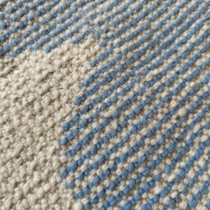 Pu&ograve; includere: Primo piano di un tappeto strutturato con sfondo color crema e strisce diagonali in azzurro. La trama del tappeto &egrave; visibile, creando un aspetto tattile. Il design &egrave; semplice e moderno, adatto a vari stili di interni.