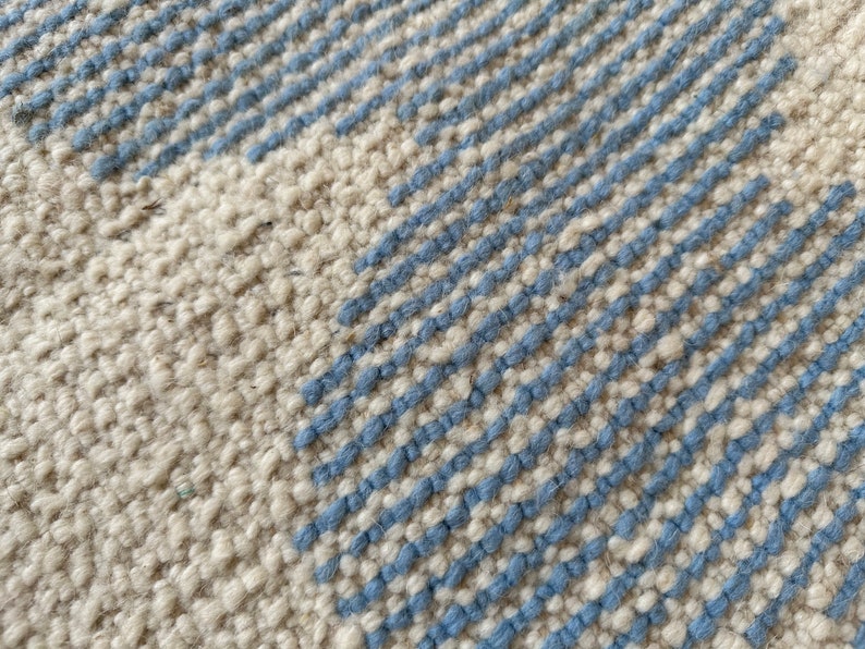 Pu&ograve; includere: Primo piano di un tappeto strutturato con sfondo color crema e strisce diagonali in azzurro. La trama del tappeto &egrave; visibile, creando un aspetto tattile. Il design &egrave; semplice e moderno, adatto a vari stili di interni.