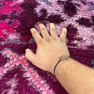 Pu&ograve; includere: Una mano &egrave; appoggiata su un tappeto morbido e dai colori vivaci. Il tappeto presenta un mix di tonalit&agrave; rosa, viola e bordeaux in un motivo strutturato. Un braccialetto con un ciondolo decorativo &egrave; sul polso.