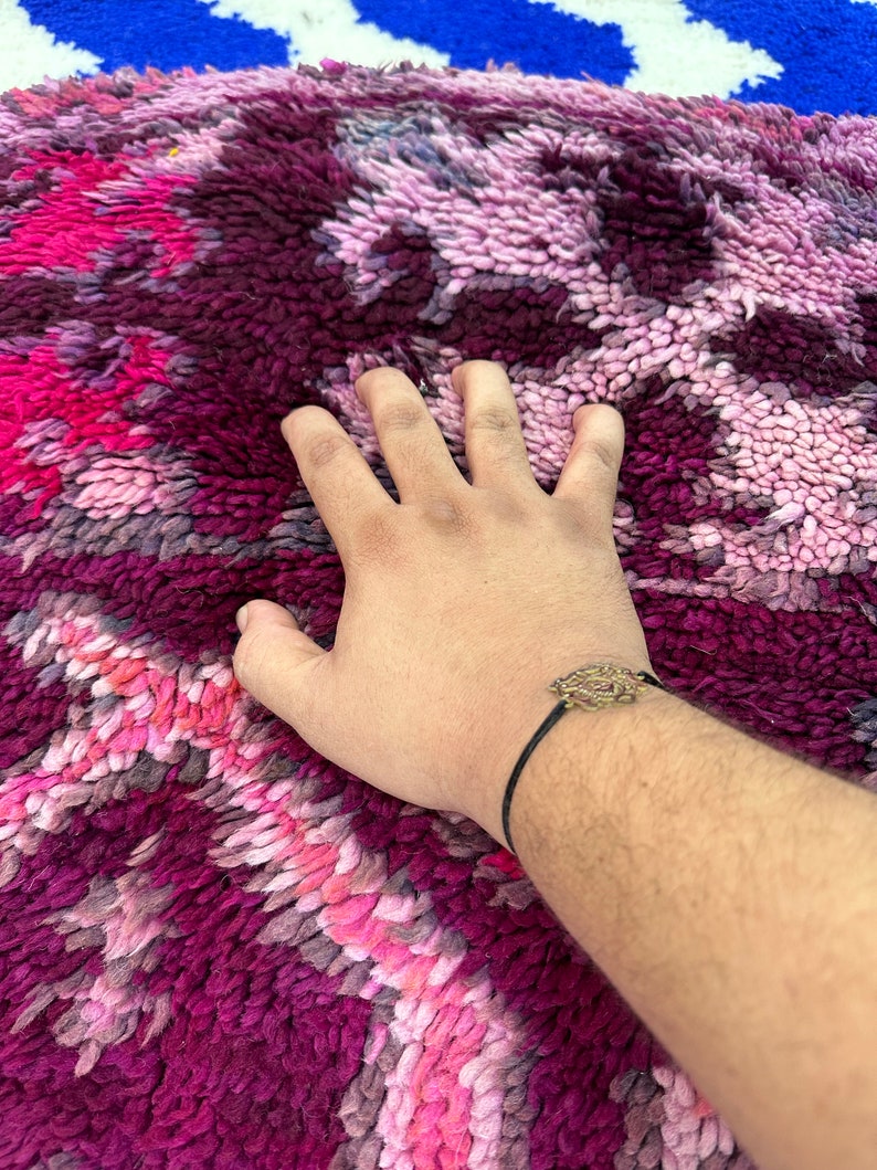 Pu&ograve; includere: Una mano &egrave; appoggiata su un tappeto morbido e dai colori vivaci. Il tappeto presenta un mix di tonalit&agrave; rosa, viola e bordeaux in un motivo strutturato. Un braccialetto con un ciondolo decorativo &egrave; sul polso.