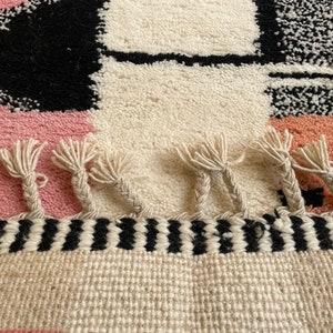 May include: A hand-woven rug with a geometric pattern in black, white, pink, and peach. The rug has a fringe border with a black and white striped band.