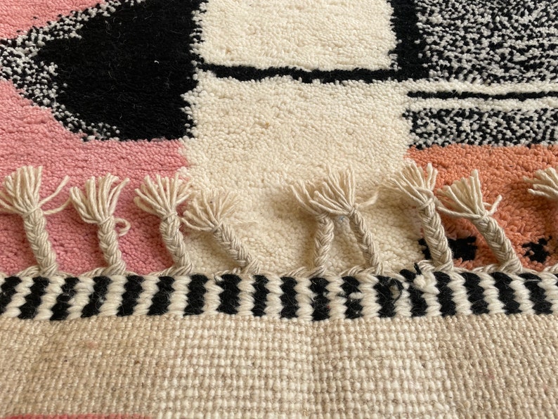 May include: A hand-woven rug with a geometric pattern in black, white, pink, and peach. The rug has a fringe border with a black and white striped band.