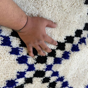 May include: White, black, and blue Moroccan rug with a geometric pattern. The rug is made of wool and has a soft, fluffy texture.