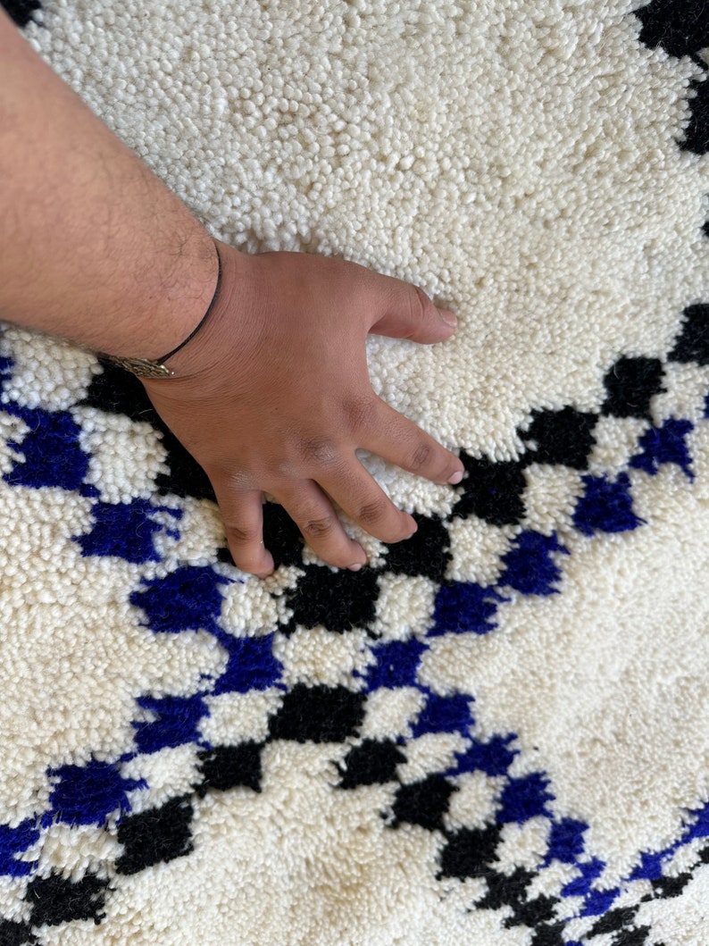 May include: White, black, and blue Moroccan rug with a geometric pattern. The rug is made of wool and has a soft, fluffy texture.